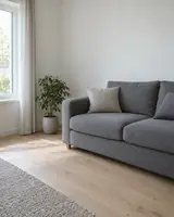 A plain living room corner with a grey sofa against a bare white wall, a potted plant and a pale wooden floor