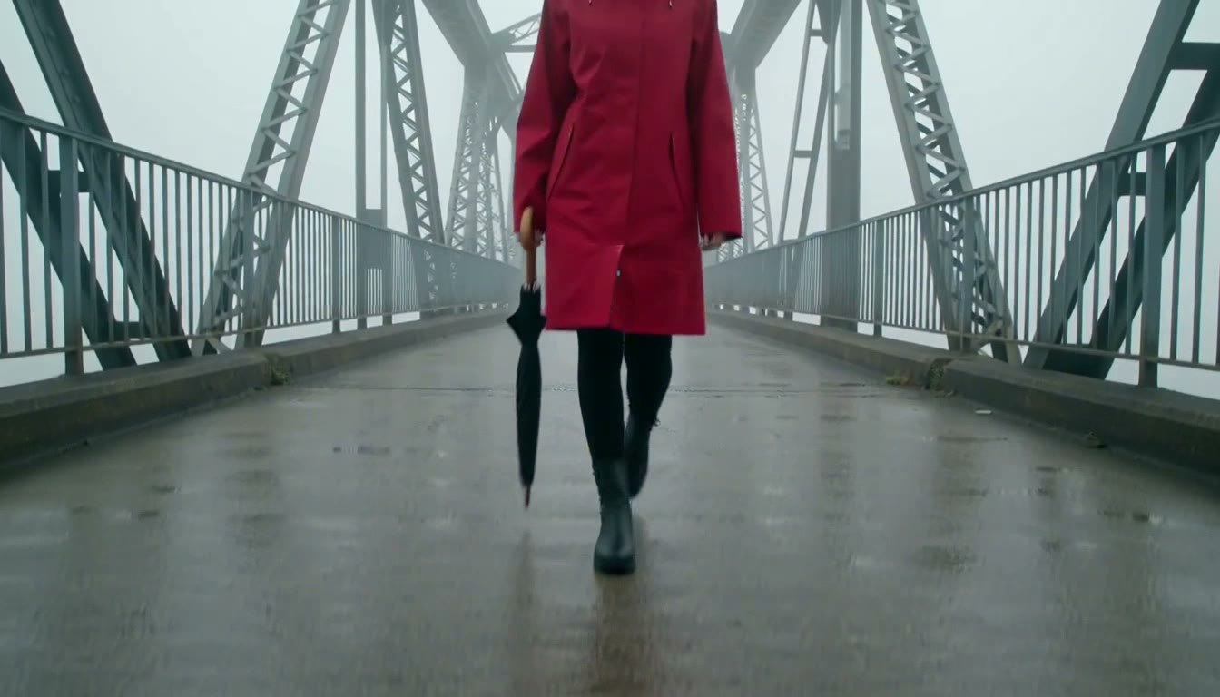 A woman in a red raincoat walking across a bridge in fog, carrying a closed umbrella, a clip made with MiniMax H3 Max